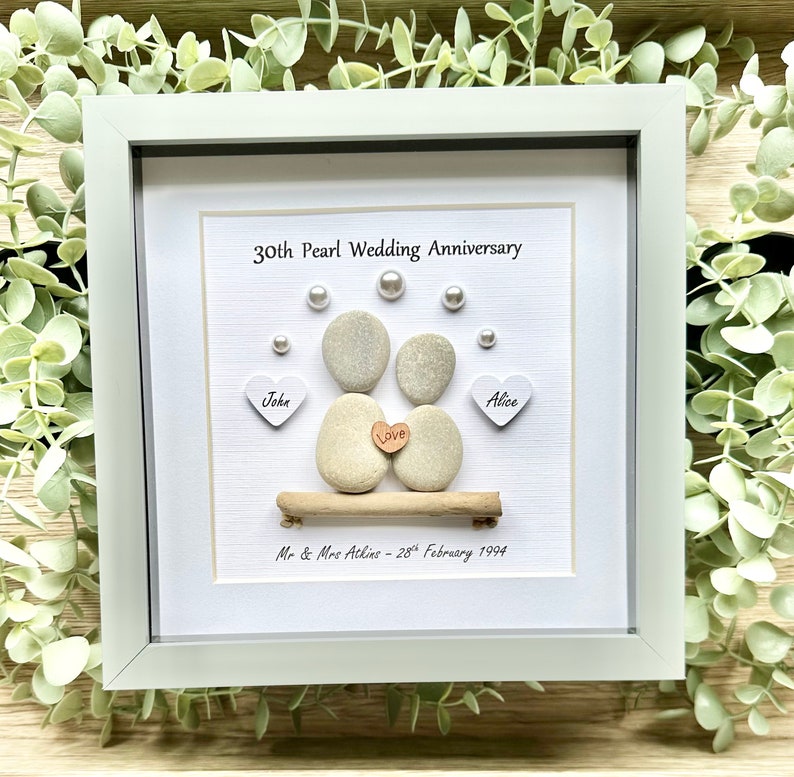 May include: A white framed picture with a 30th Pearl Wedding Anniversary message. The picture features two gray stones representing a couple, with a small wooden heart that says "Love" between them. The stones are sitting on a small piece of driftwood. The names "John" and "Alice" are written on small white hearts below the stones. The date "28th February 1994" is written below the driftwood.