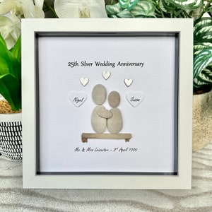 May include: A white framed picture with a white background. The picture features a pebble art design of a couple holding each other. The text "25th Silver Wedding Anniversary" is at the top of the picture. The names "Nigel" and "Susan" are written on hearts on either side of the couple. The text "Mr & Mrs Leicester - 3rd April 1999" is at the bottom of the picture.