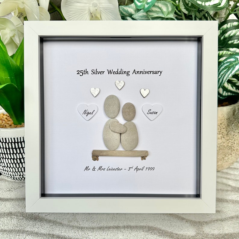 May include: A white framed picture with a white background. The picture features a pebble art design of a couple holding each other. The text "25th Silver Wedding Anniversary" is at the top of the picture. The names "Nigel" and "Susan" are written on hearts on either side of the couple. The text "Mr & Mrs Leicester - 3rd April 1999" is at the bottom of the picture.