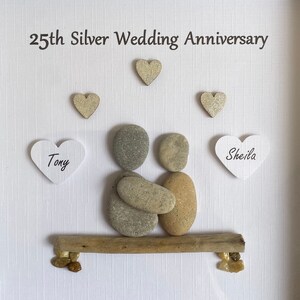 May include: A 25th Silver Wedding Anniversary pebble art piece featuring a couple sitting on a driftwood bench. The couple is made of gray and brown pebbles, and the bench is made of driftwood. The names "Tony" and "Sheila" are written on white hearts.