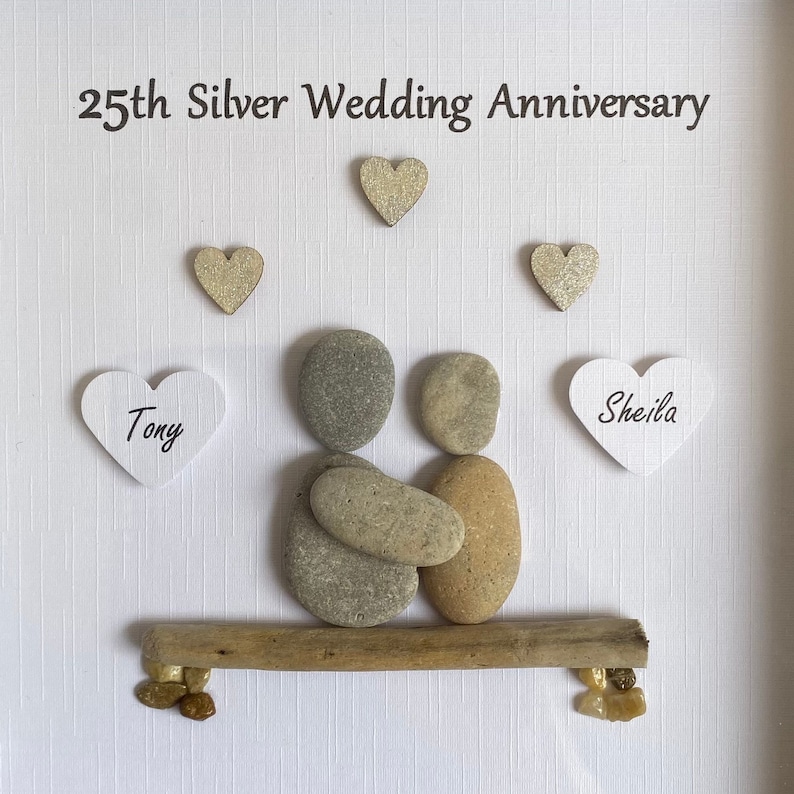 May include: A 25th Silver Wedding Anniversary pebble art piece featuring a couple sitting on a driftwood bench. The couple is made of gray and brown pebbles, and the bench is made of driftwood. The names "Tony" and "Sheila" are written on white hearts.