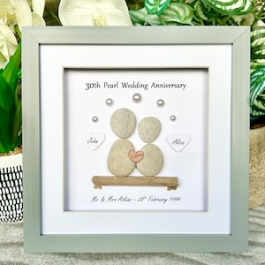 May include: A framed pebble art piece celebrating a 30th wedding anniversary. The art features two gray pebbles representing a couple, with the names "John" and "Alice" written on white hearts. The pebbles are holding a small heart with the word "Love" written on it. The text "30th Pearl Wedding Anniversary" and "Mr & Mrs Atkins - 28th February 1994" are also included.