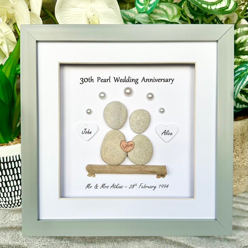 May include: A framed pebble art piece celebrating a 30th wedding anniversary. The art features two gray pebbles representing a couple, with the names "John" and "Alice" written on white hearts. The pebbles are holding a small heart with the word "Love" written on it. The text "30th Pearl Wedding Anniversary" and "Mr & Mrs Atkins - 28th February 1994" are also included.