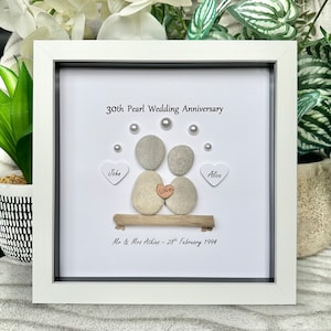 May include: A white framed picture with a white background. The picture is a pebble art piece of a couple sitting on a driftwood log. The couple is made of gray and brown pebbles. The picture is decorated with white pearls and a small wooden heart that says "Love". The text on the picture reads "30th Pearl Wedding Anniversary", "John", "Alice", "Mr & Mrs Atkins - 28th February 1994".