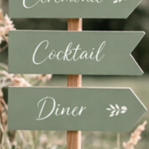 Peut inclure: Panneau directionnel en bois avec trois flèches vertes indiquant "Cérémonie", "Cocktail" et "Diner" en écriture blanche. Le panneau est planté dans un cadre naturel avec de la verdure.