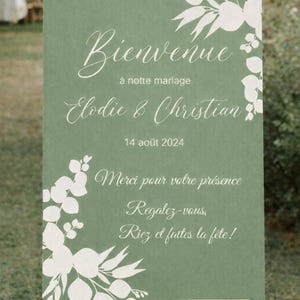 May include: A green wedding welcome sign on a wooden easel. The sign has white floral accents and elegant script that reads "Bienvenue" and the names "Elodie & Christian" with the date "14 ao&ucirc;t 2024".