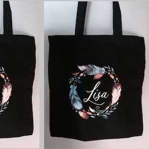 Peut inclure: Sac fourre-tout noir avec une couronne de plumes aquarelle dans les tons de bleu, rose et marron. Un sac porte le texte "EVJF de Géraldine" et l'autre le nom "Lisa" en cursive.