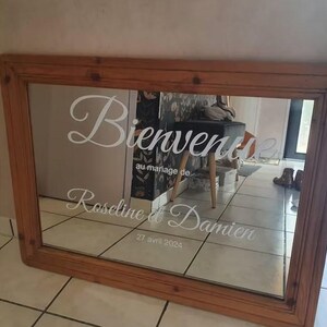 May include: A rectangular wooden framed mirror with the words "Bienvenue" and "au mariage de Roseline et Damien 27 avril 2024" in white script. The mirror reflects a room.