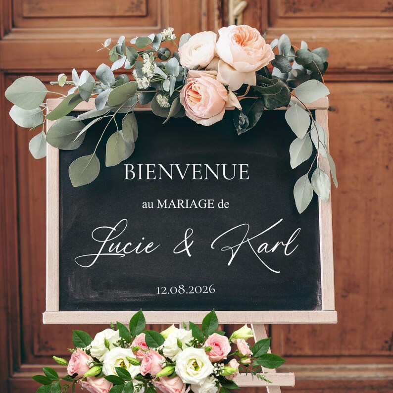 May include: A chalkboard welcome sign for a wedding, with the words "BIENVENUE au MARIAGE de Lucie & Karl 12.08.2026" in white script. The sign is decorated with pink and white roses, eucalyptus, and greenery. The sign is on a wooden easel.