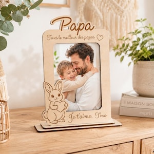 Peut inclure: Un cadre photo en bois clair avec une découpe de lapin tenant un cœur en bas à gauche. Le haut est gravé du mot Papa et du texte Tu es le meilleur des papas, tandis que le bas porte l'inscription Je t'aime. Tino.