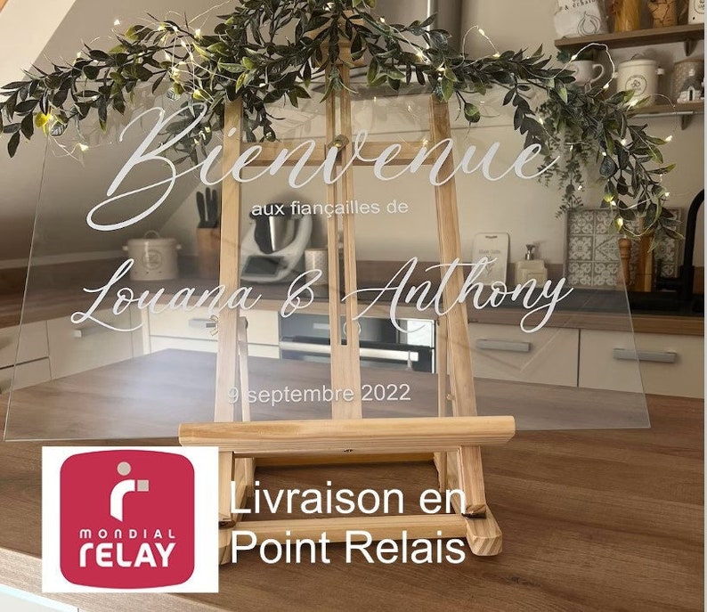 May include: A clear acrylic welcome sign on a wooden easel, with the words "Bienvenue" and the names "Louana & Anthony" in elegant white script. The date "9 septembre 2022" is also displayed. The sign is decorated with greenery and fairy lights.