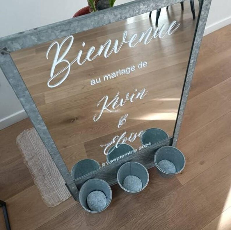 May include: A large, rectangular mirror with a metal frame. The mirror has white cursive text that reads "Bienvenue au mariage de Kevin & Elo&iuml;se 21 septembre 2024". Below the mirror are four small, gray metal buckets.