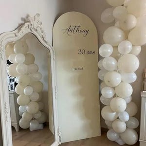 May include: A cream-colored arch-shaped sign with the name "Anthony" and the text "30 ans" and "jan 2024" in black script. White balloons are arranged on either side of the sign and in front of a large ornate mirror.