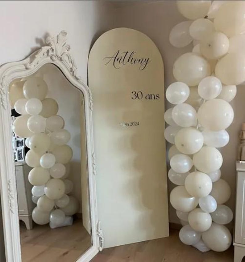 May include: A cream-colored arch-shaped sign with the name "Anthony" and the text "30 ans" and "jan 2024". The sign is flanked by white balloons and a decorative mirror.
