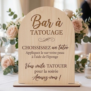 Peut inclure: Un panneau en bois clair en forme d'arche avec un texte gravé en brun foncé : Bar à TATOUAGE, CHOISSISSEZ un tatoo, Appliquez le sur votre peau à l'aide de l'éponge, Vous voilà TATOUER pour la soirée, Amusez vous ! Le panneau repose sur un socle.