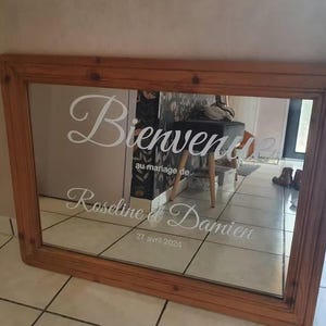 May include: A rectangular wooden framed mirror with the words "Bienvenue" and "au mariage de Roseline et Damien 27 avril 2024" in white script. The mirror reflects a room.