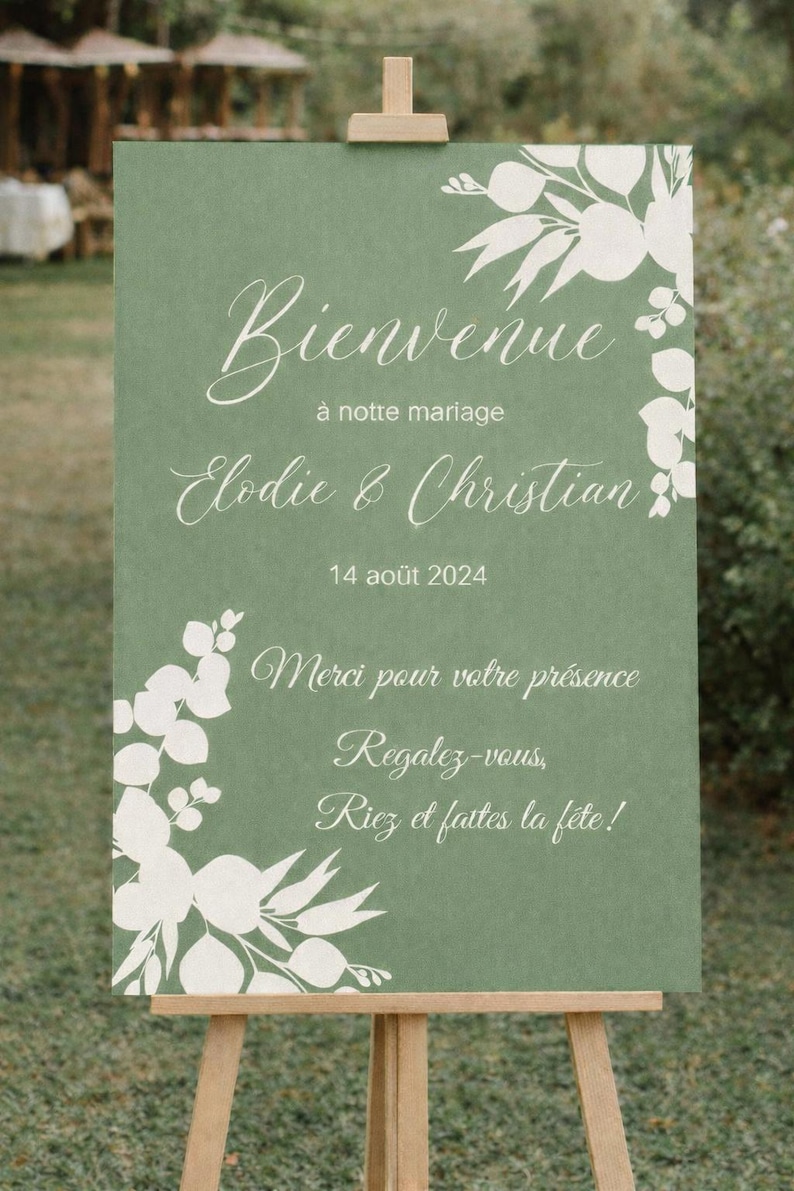 May include: A green wedding welcome sign on a wooden easel. The sign has white floral accents and elegant script that reads "Bienvenue" and the names "Elodie & Christian" with the date "14 ao&ucirc;t 2024".