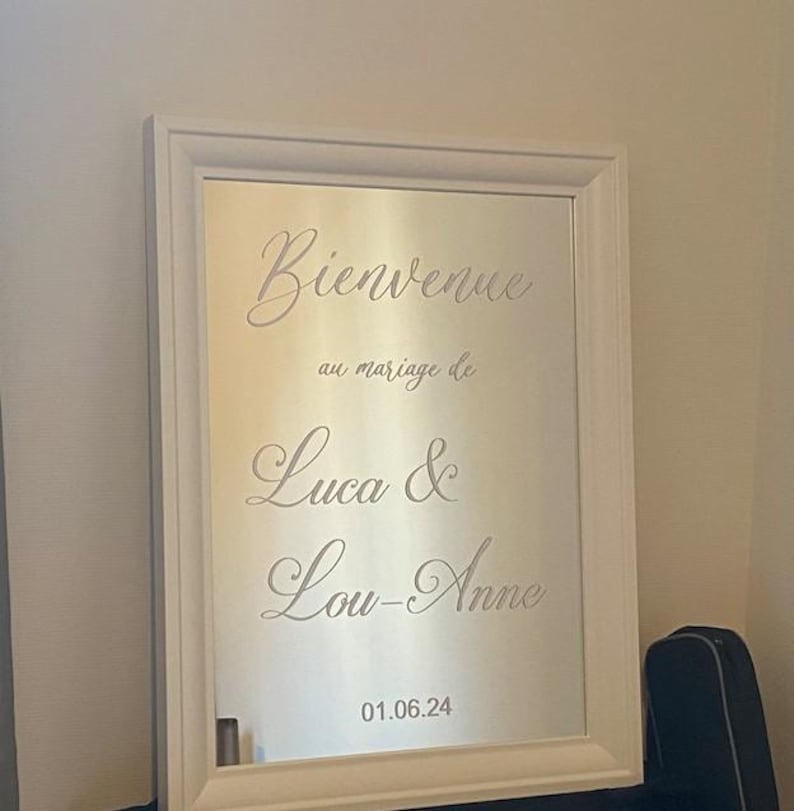 May include: A white framed mirror with the words "Bienvenue au mariage de Luca & Lou-Anne" in elegant script. The date "01.06.24" is displayed at the bottom. The mirror is set against a neutral background.