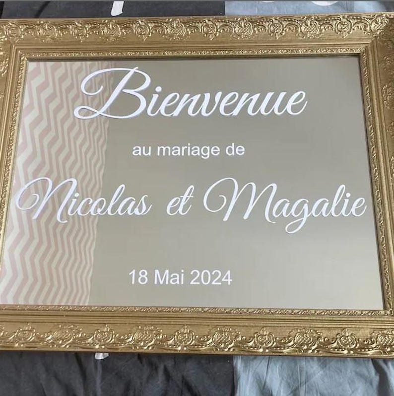 May include: A gold-framed mirror with the words "Bienvenue" and "Nicolas et Magalie" in white script. The date "18 Mai 2024" is also displayed. The frame has an ornate, decorative design, suitable for a wedding.