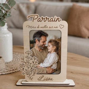 Peut inclure: Un cadre photo en bois clair gravé avec le texte Parrain, Merci de veiller sur moi et Lison. Le cadre présente un motif floral linéaire décoratif sur le côté gauche et contient une photographie montrant un adulte et un enfant.