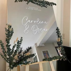 May include: A large, reflective wedding sign with the names "Caroline & Oscar" in elegant white script. The sign is decorated with greenery and the date "31.08.2024". The text "Bienvenue A Notre Mariage" is at the top.