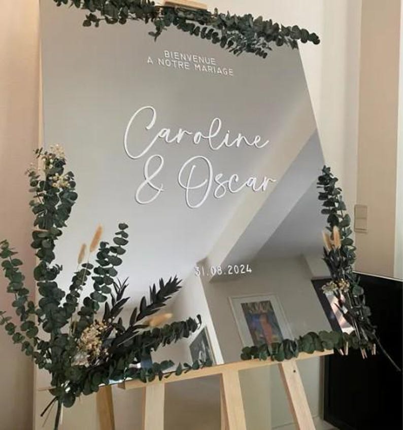 May include: A large, reflective wedding sign with the names "Caroline & Oscar" in elegant white script. The sign is decorated with greenery and the date "31.08.2024". The text "Bienvenue A Notre Mariage" is at the top.
