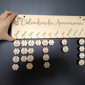 Peut inclure: Un calendrier d'anniversaires en bois clair avec l'inscription "Calendrier des Anniversaires" gravée en haut. Il comporte douze colonnes pour les mois, avec des anneaux métalliques retenant des disques et des hexagones en bois.