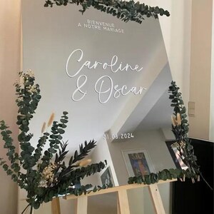 May include: A large, reflective wedding sign with the names "Caroline & Oscar" in elegant white script. The sign is decorated with greenery and the date "31.08.2024". The text "Bienvenue A Notre Mariage" is at the top.