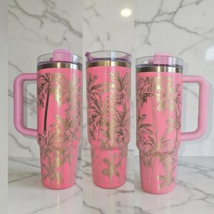 May include: Three pink insulated tumblers with gold palm tree designs and pink handles. The tumblers have clear lids and the brand name "STANLEY" is visible on the side. The tumblers are designed to keep drinks cold or hot.