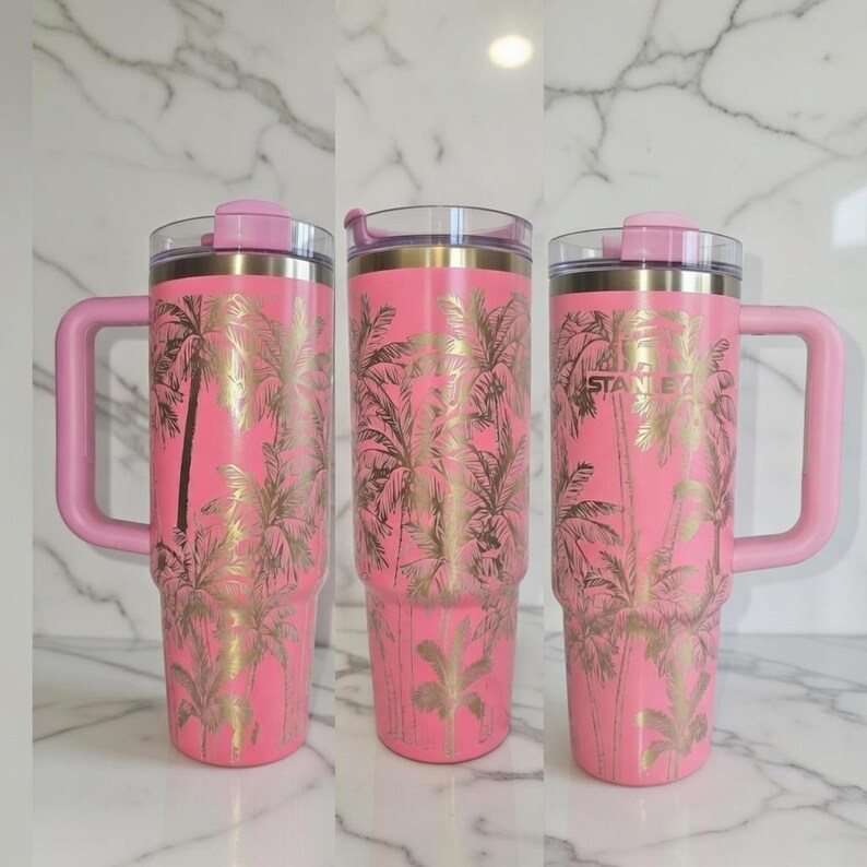 May include: Three pink insulated tumblers with gold palm tree designs and pink handles. The tumblers have clear lids and the brand name "STANLEY" is visible on the side. The tumblers are designed to keep drinks cold or hot.