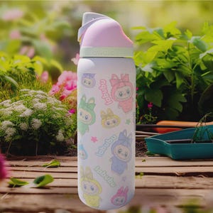 Puede incluir: Una botella de agua blanca Owala con personajes de dibujos animados coloridos y la palabra "labubu". La botella tiene una tapa rosa y gris con un asa. El fondo muestra un jard&iacute;n con plantas verdes y flores rosas.