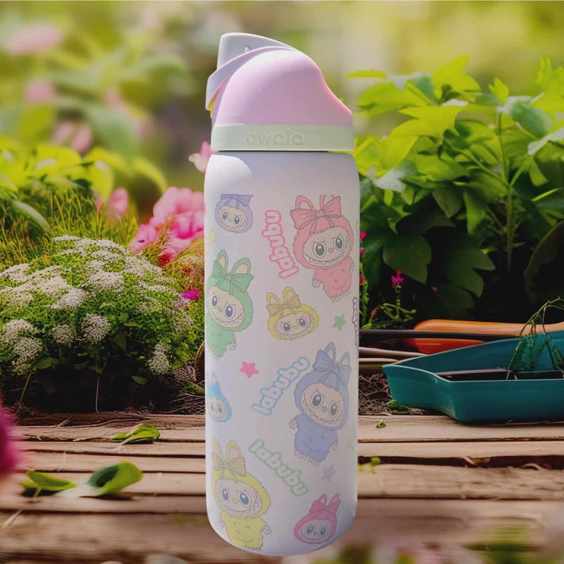 Puede incluir: Una botella de agua blanca Owala con personajes de dibujos animados coloridos y la palabra "labubu". La botella tiene una tapa rosa y gris con un asa. El fondo muestra un jard&iacute;n con plantas verdes y flores rosas.