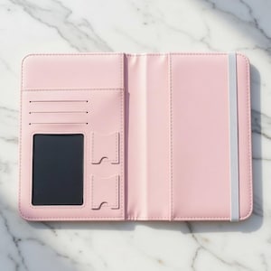 Puede incluir: Funda de cuaderno de polipiel rosa claro abierta, con ranuras para tarjetas, una ventana de identificaci&oacute;n transparente y un cierre de banda el&aacute;stica. El cuaderno est&aacute; abierto, revelando las p&aacute;ginas interiores. La funda est&aacute; sobre una superficie de m&aacute;rmol blanco.