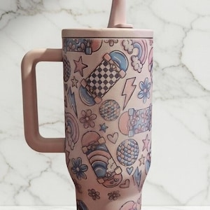 May include: A pink tumbler with a handle, lid, and straw, adorned with a repeating pattern of skateboards, stars, flowers, rainbows, and lightning bolts. The tumbler is designed to hold approximately 30 ounces.