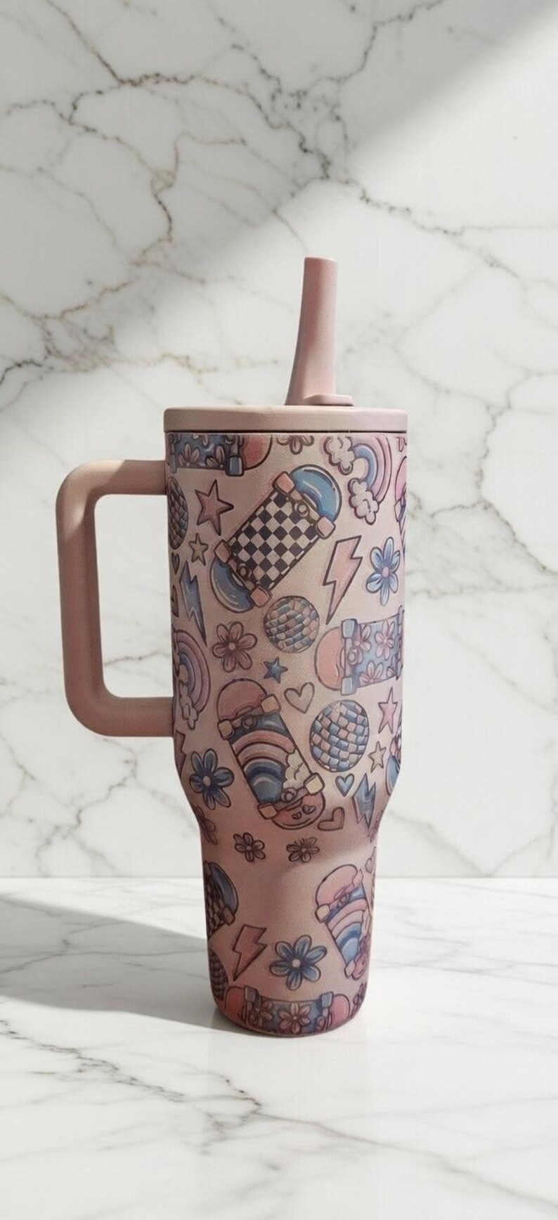 May include: A pink tumbler with a handle, lid, and straw, adorned with a repeating pattern of skateboards, stars, flowers, rainbows, and lightning bolts. The tumbler is designed to hold approximately 30 ounces.