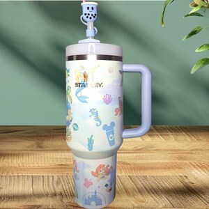 May include: A blue and white stainless steel travel mug with a lid and handle. The mug has a cartoon design featuring a mermaid, seahorses, and other sea creatures. The mug also has the text "STANLEY" on it.