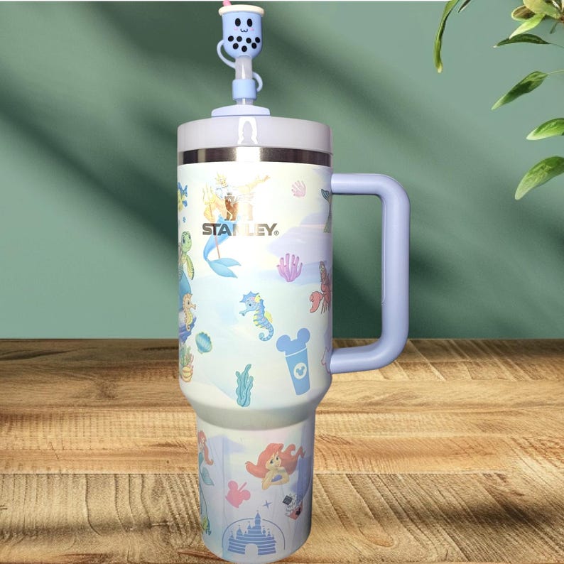 May include: A blue and white stainless steel travel mug with a lid and handle. The mug has a cartoon design featuring a mermaid, seahorses, and other sea creatures. The mug also has the text "STANLEY" on it.