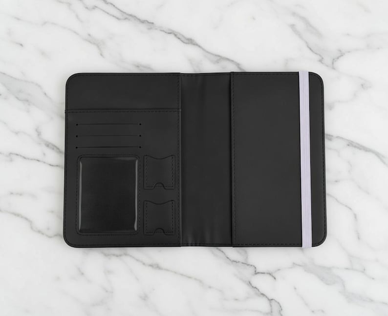 Puede incluir: Portapasaporte negro abierto con cierre de banda el&aacute;stica blanca. El interior presenta ranuras para tarjetas, una ventana de identificaci&oacute;n transparente y dos bolsillos adicionales. Sobre una superficie de m&aacute;rmol.