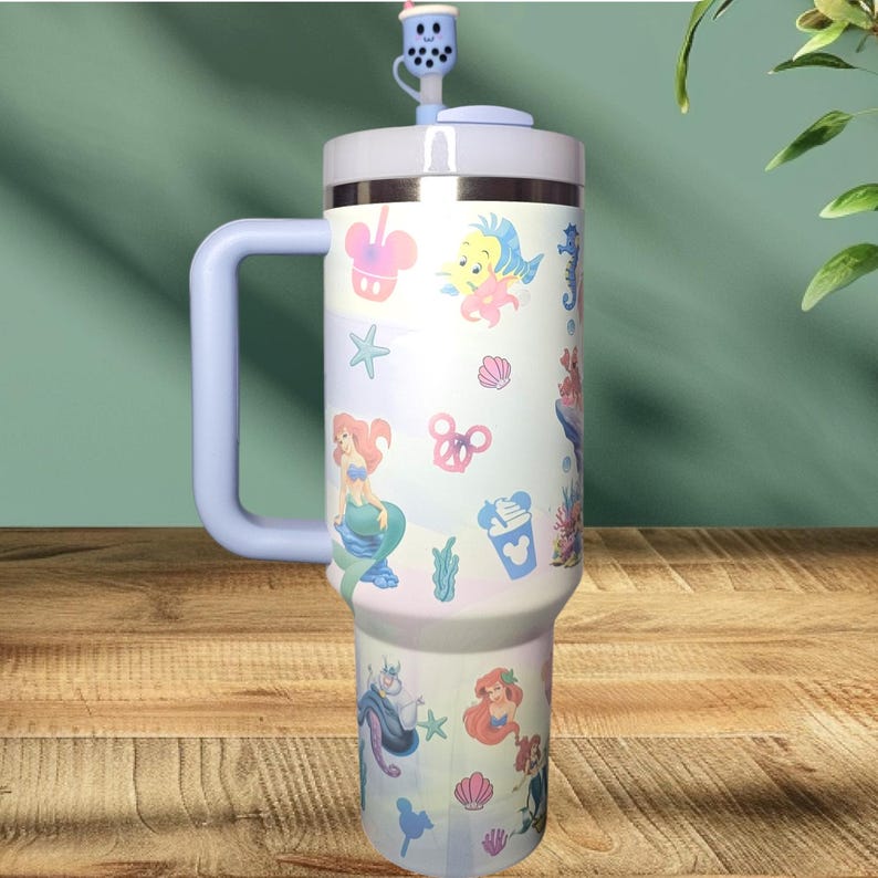 May include: A light blue stainless steel tumbler with a handle and a straw. The tumbler has a white background with a variety of colorful cartoon images of Disney characters from The Little Mermaid, including Ariel, Ursula, Sebastian, and Flounder. The tumbler also has a few Mickey Mouse ears and seashells.