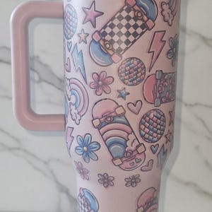 May include: Pink travel mug with a handle, covered in a pattern of skateboards, stars, flowers, rainbows, and lightning bolts. The mug has a matching pink lid and handle. The design is in shades of pink, blue, and white.