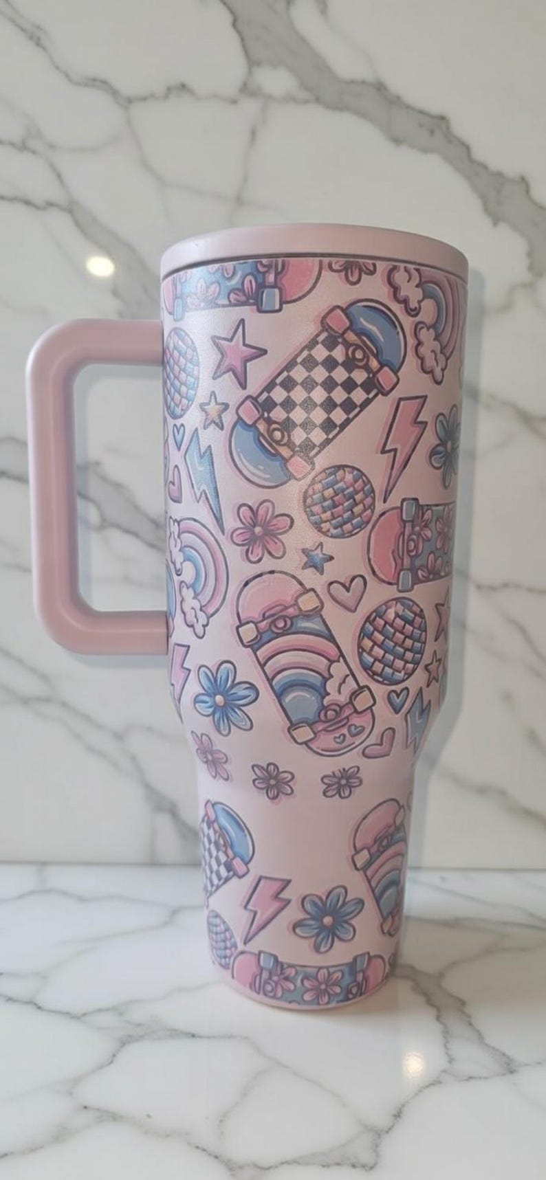 May include: Pink travel mug with a handle, covered in a pattern of skateboards, stars, flowers, rainbows, and lightning bolts. The mug has a matching pink lid and handle. The design is in shades of pink, blue, and white.