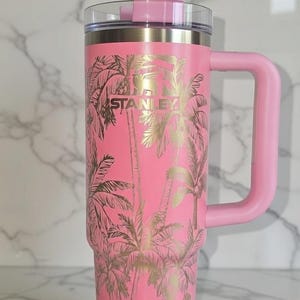 May include: Pink insulated tumbler with a handle, featuring a gold palm tree design. The tumbler has a clear lid and the brand name "STANLEY" is visible. A stylish drinkware item for hot or cold beverages.