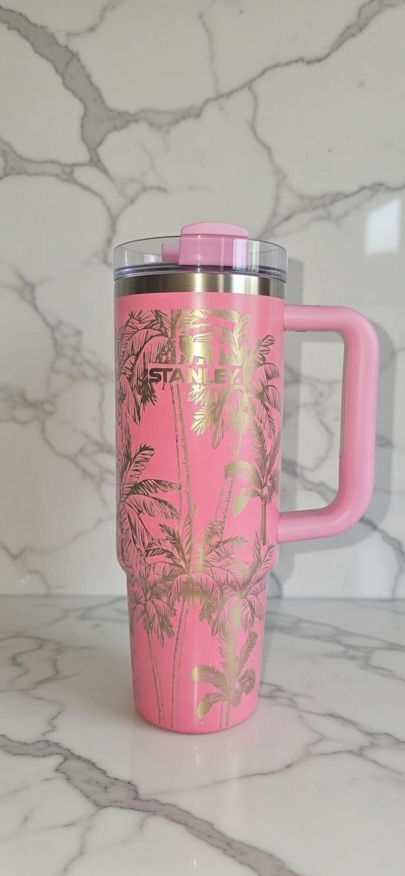 May include: Pink insulated tumbler with a handle, featuring a gold palm tree design. The tumbler has a clear lid and the brand name "STANLEY" is visible. A stylish drinkware item for hot or cold beverages.