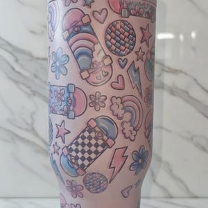May include: A pink tumbler with a repeating pattern of skateboards, flowers, hearts, stars, lightning bolts, rainbows, and disco balls. The tumbler is approximately 10 inches tall.