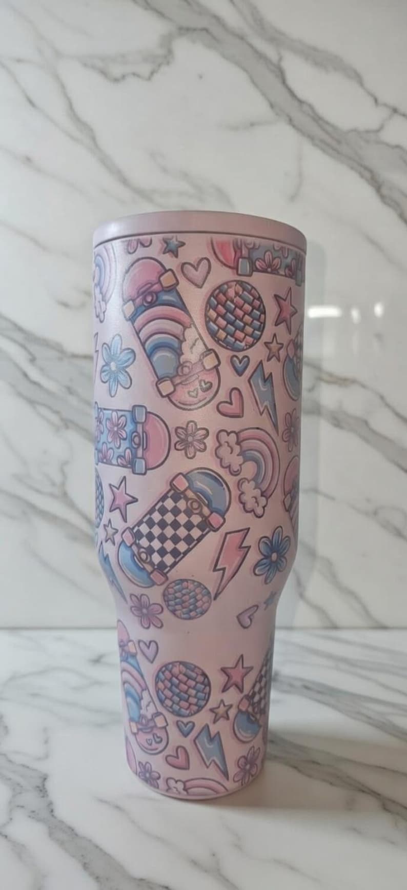 May include: A pink tumbler with a repeating pattern of skateboards, flowers, hearts, stars, lightning bolts, rainbows, and disco balls. The tumbler is approximately 10 inches tall.