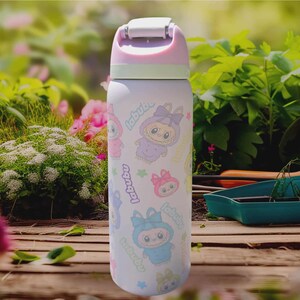 Puede incluir: Una botella de agua de color lila claro con tapa abatible, con un patr&oacute;n repetido de personajes de dibujos animados y la palabra "labubu". La tapa es rosa y verde. La botella es cil&iacute;ndrica y parece ser de metal.