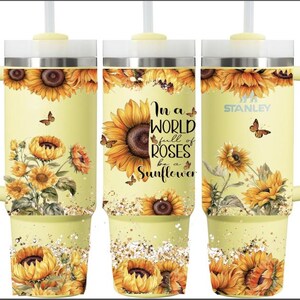 Puede incluir: Un vaso Stanley amarillo con un dise&ntilde;o de girasoles y mariposas, y el texto "In a world full of roses be a sunflower". El vaso tiene un asa y una pajita.