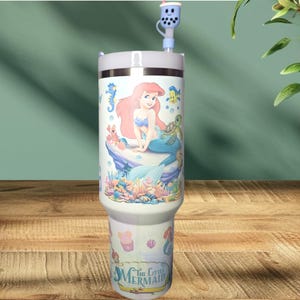 May include: A white and blue stainless steel tumbler with a lid and straw. The tumbler features a colorful image of Ariel, the Little Mermaid, with a seahorse, a crab, and a turtle. The text "The Little Mermaid" is printed on the bottom of the tumbler.