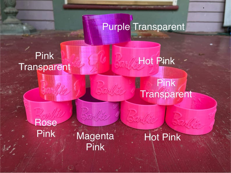 May include: A stack of eight colorful plastic rings with the word "Barbie" embossed on each ring. The rings are in shades of pink, magenta, and purple. The colors are labeled as "Purple Transparent", "Hot Pink", "Pink Transparent", "Rose Pink", "Magenta Pink", and "Hot Pink".