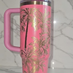 May include: Pink insulated tumbler with a handle, featuring a gold palm tree design. The tumbler has a pink lid and handle. The tumbler is approximately 10 inches tall.